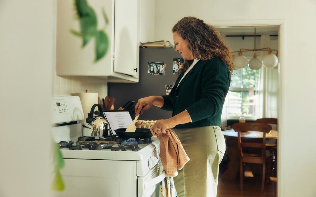 Mulher cozinhando no fogão da cozinha de casa, com o notebook aberto ao lado