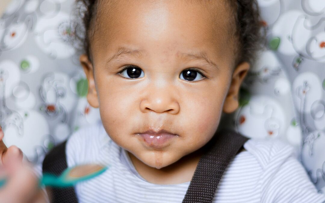 Bebê de pele morena na cadeirinha, olhando para a câmera enquanto recebe comida na colher